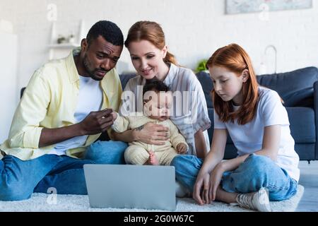 famiglia interracial seduto sul pavimento e guardando film sul computer portatile Foto Stock