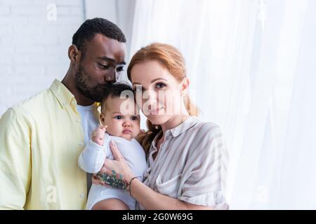 donna felice guardando la macchina fotografica vicino sorridente marito baciando la testa della figlia infantile Foto Stock