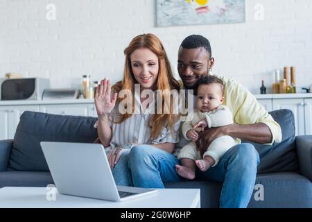 donna sorridente che agita la mano vicino al computer portatile e alla famiglia interracial Foto Stock