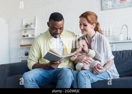 libro di lettura dell'uomo afroamericano vicino alla moglie sorridente e alla figlia infantile Foto Stock