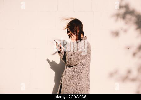 Vista laterale di una donna felice che usa lo smartphone mentre si trova in piedi contro la parete Foto Stock