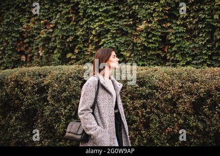 Giovane donna che cammina in città contro piante verdi Foto Stock