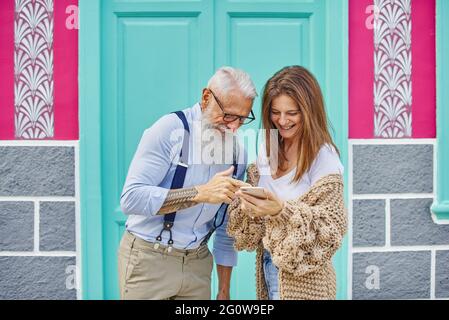 Felice donna di mezza età che condivide i dati nei social media con un elegante uomo anziano mentre naviga sul telefono cellulare in strada contro un edificio colorato Foto Stock
