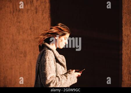 Vista laterale di una donna non binaria che usa uno smartphone che cammina contro la parete Foto Stock