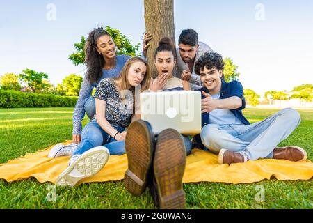 Gruppo di cinque giovani felici e diverse persone multirazziali di generazione z seduti sull'erba in un campo di parco cittadino utilizzando il computer portatile per navigare in rete. Donna scioccata con s Foto Stock
