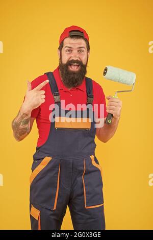 caposquadra che sta andando fare la parete che affronta. uomo brutale felice in uniforme del boilersuit. hipster maturo usa l'attrezzo del rullo della vernice per la pittura. riparatore o costruttore Foto Stock