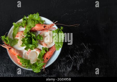 Capesante grezzo naturale con gamberi bolliti nel lavandino in una ciotola di ceramica con foglie di lattuga su sfondo scuro. Messa a fuoco selettiva. Foto Stock