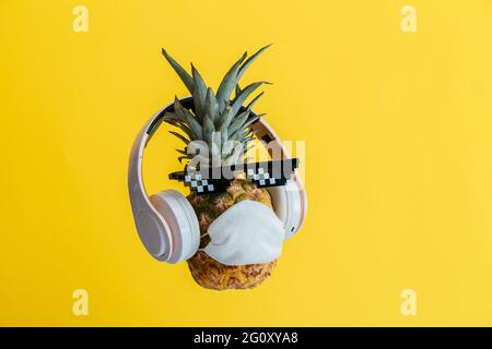 Creativo divertente ananas viso indossare occhiali cuffie e maschera medica di protezione. Riposante faccia di ananas levitating sul colore giallo estate Foto Stock