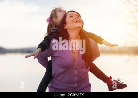 Una mamma giocosa che dà la figlia piggy indietro al parco estivo. Sia sorridente che guardando in su. Primavera, estate parco vicino lago background.Closeup. Foto Stock