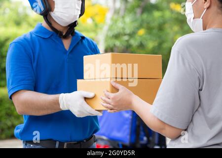 Uomo di consegna asiatico che indossa maschera facciale e uniforme blu con moto che consegna il servizio espresso della scatola del pacco alla cliente della donna Foto Stock