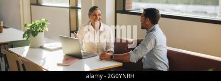 donna d'affari che sembra felice durante una riunione con un collega alla scrivania dell'ufficio. Donna sorridente che indica il notebook e parla con un collega in ufficio. Foto Stock