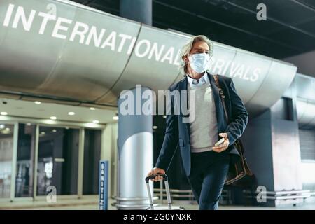 Uomo d'affari che indossa una maschera protettiva con i bagagli che camminano all'interno del terminal dell'aeroporto. Uomo che viaggia durante l'epidemia di coronavirus. Foto Stock