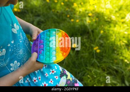 La ragazza gioca con il tocco anti-stress silicone pop esso giocattolo all'aperto in giardino. Le dita premono le bolle e i pulsanti morbidi dell'arcobaleno. Gioco di tendenza per i deve Foto Stock