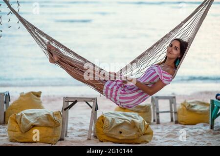 Resort. Una giovane, abbronzata, bella donna in un abito a righe è sdraiata su un'amaca in vimini. Vista laterale. Sullo sfondo, il mare o l'oceano. Vacanze in Foto Stock