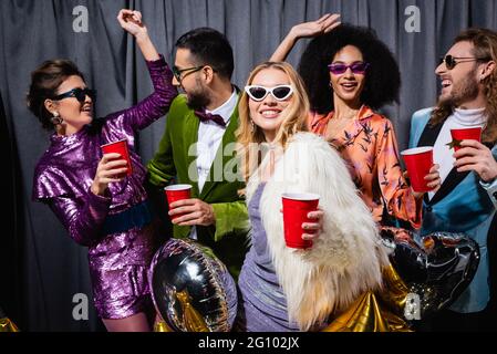 divertenti amici interrazziali in abiti colorati con tazze di plastica in mani che ballano su sfondo grigio Foto Stock