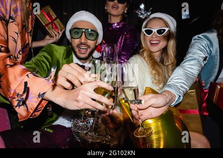 sorridenti amici interrazziali in cappelli di santa e occhiali da sole che si aggrappano con bicchieri di champagne su sfondo nero Foto Stock