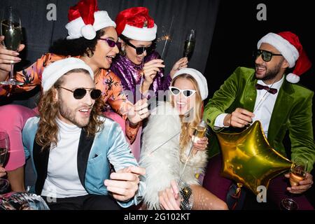 amici interrazziali a santa cappelli bere champagne e celebrare il nuovo anno vicino tenda grigia su sfondo nero Foto Stock