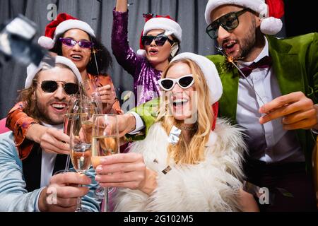 sorridenti amici interrazziali in santa cappelli celebrare il nuovo anno vicino tenda grigio su sfondo nero Foto Stock