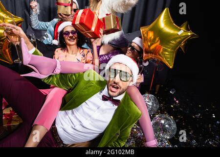 giocosi amici interrazziali in cappelli di santa e occhiali da sole che celebrano il nuovo anno vicino tenda grigia su sfondo nero Foto Stock