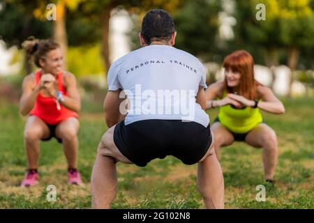Allenatore di fitness maschile nel parco con due donne felici e facendo squat. Foto Stock