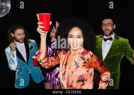 donna afroamericana positiva con gesto di tostatura sulla festa con amici multirazziali su sfondo nero Foto Stock