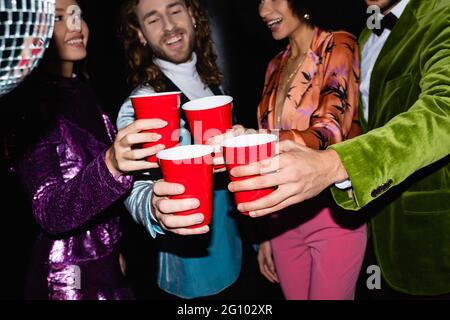 amici multirazziali positivi in abiti colorati che tostano con tazze di plastica nel night club su sfondo nero Foto Stock