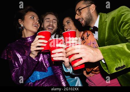 amici multirazziali positivi in abiti colorati che tostano con tazze di plastica su sfondo nero Foto Stock