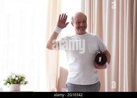Uomo anziano che tiene il tappeto yoga che si allena dicendo ciao felice e sorridente Foto Stock