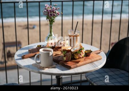 Sandwich di pollo e caffè ghiacciato con cacao caldo e fiore in vaso su tavolo di marmo nel caffè vicino alla spiaggia al mattino Foto Stock