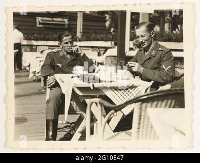Luftwaffe soldati su una terrazza. Tre soldati Luftwaffe sono seduti su una terrazza. Sul tavolo sono tazze e bugie che cosa assomiglia ad una borsa per una macchina fotografica. Solo il braccio può essere visto dal terzo soldato. Entrambi gli altri sono barehead e fumare una sigaretta. A sinistra è ancora il cameriere da vedere. Foto Stock