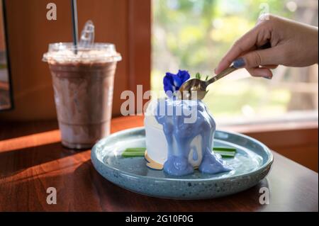 Mano della donna che usa un cucchiaio per tagliare la torta di cocco con la salsa di piselli a farfalla su un piatto di ceramica e cacao ghiacciato sul tavolo nel caffè Foto Stock