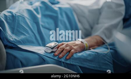 Hospital Ward: Donna anziana che riposa in un letto con pulsossimetro Finger Heart Rate Monitor che mostra il polso. Le sue mani fragili poggiano su una coperta Foto Stock