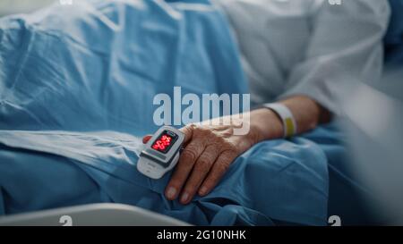 Hospital Ward: Donna anziana che riposa in un letto con pulsossimetro Finger Heart Rate Monitor che mostra il polso. Le sue mani fragili poggiano su una coperta Foto Stock