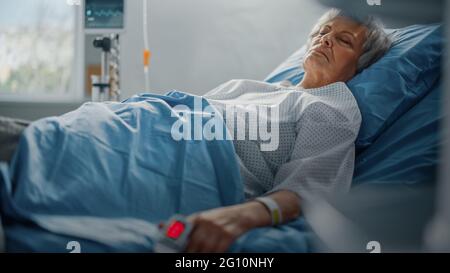 Hospital Ward: Donna anziana che riposa in un letto con pulsossimetro Finger Heart Rate Monitor che mostra il polso. Le sue mani fragili poggiano su una coperta Foto Stock