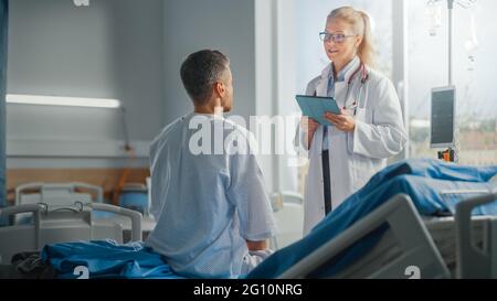 Hospital Ward: Seduta sul letto paziente maschio caucasico ascolta il medico femminile esperto che spiega i risultati dei test, dà consigli, usato Tablet computer Foto Stock