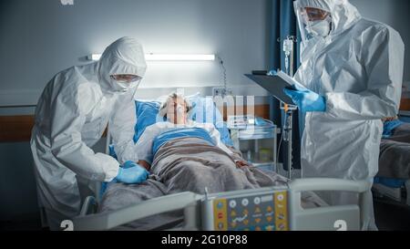 Hospital Coronavirus Emergency Department Ward: Medico che indossa tuta scrive i segni vitali di un paziente femminile anziano con la maschera di ossigeno che riposa nel letto Foto Stock