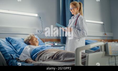 Hospital Ward: Il medico amichevole chiede assistenza sanitaria Domande di sorridere anziana Signora che riposa a letto. Il medico usa Tablet computer, fa Checkup, parla Foto Stock
