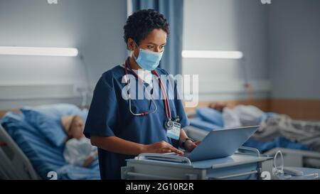 Hospital Ward: Professionista nero latino femmina testa infermiere o medico indossare maschera viso utilizza un computer medico. Nella sezione pazienti in background in Letti Foto Stock