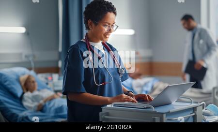 Hospital Ward: Professionista sorridente nero femmina testa infermiere o medico indossando stetoscopio utilizza un computer medico. Nella sezione pazienti in background in Letti Foto Stock