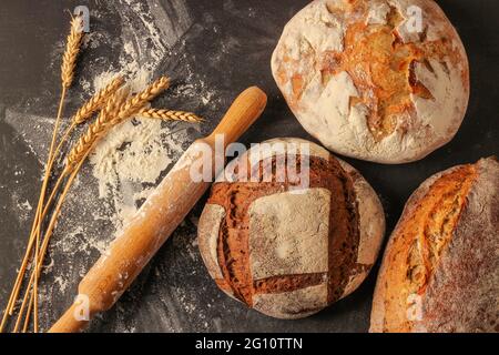 Pani di pane di grano e segale, farina, spighe di grano e un tondino di legno su sfondo nero. Vista dall'alto Foto Stock