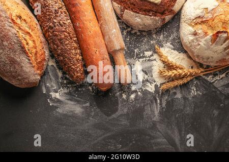 Pani di pane di grano e segale, farina, spighe di grano e un tondino di legno su sfondo nero. Vista dall'alto Foto Stock
