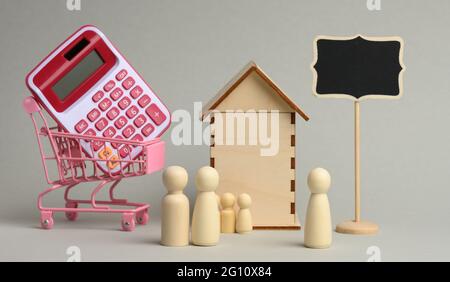 famiglia di figure in legno e un realtor sullo sfondo di una casa e una calcolatrice in un carrello su sfondo grigio. Concetto di acquisto domestico, ipoteca, Foto Stock