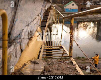 Stretto ponte tecnico con barre metalliche per la manutenzione di un grande ponte di cemento sul canale d'acqua. Ragazzo con il calcio scooter sotto il ponte. Foto Stock