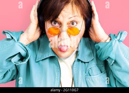 Ritratto della donna di mezza età che ascolta la musica con le cuffie - primo piano di una donna anziana con gli occhiali arancioni hippie su sfondo viola - fuoco o Foto Stock