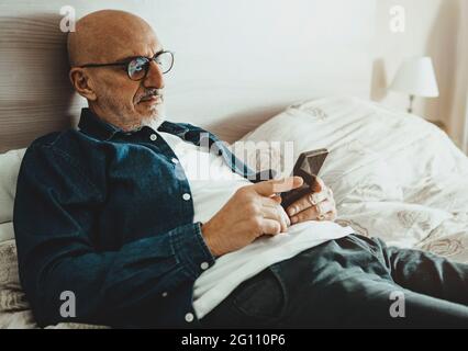 L'uomo di mezza età si appoggia sul letto con smartphone - bell'uomo legge le ultime notizie e fa shopping online dai dispositivi mobili nel tempo libero - elde Foto Stock