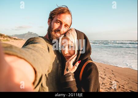 Coppia in viaggio innamorata prendendo selfie in una spiaggia deserta al tramonto - Donna con lo sguardo magnetico e il suo ragazzo hipster divertirsi guardando la camma Foto Stock