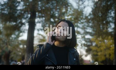 Tra alberi verdi giovane donna che cammina nel parco, parlando piacevolmente al telefono e sorrisi, gode di una conversazione con il suo amato o amico. Relazioni Foto Stock