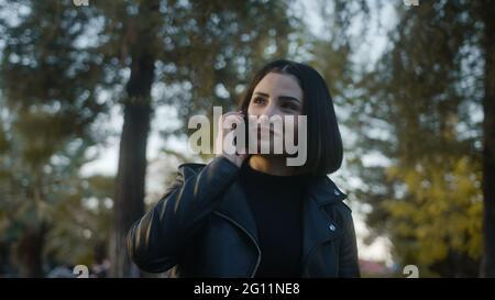 Tra alberi verdi giovane donna che cammina nel parco, parlando piacevolmente al telefono e sorrisi, gode di una conversazione con il suo amato o amico. Relazioni Foto Stock