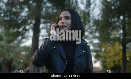 Tra alberi verdi giovane donna che cammina nel parco, parlando piacevolmente al telefono e sorrisi, gode di una conversazione con il suo amato o amico. Relazioni Foto Stock