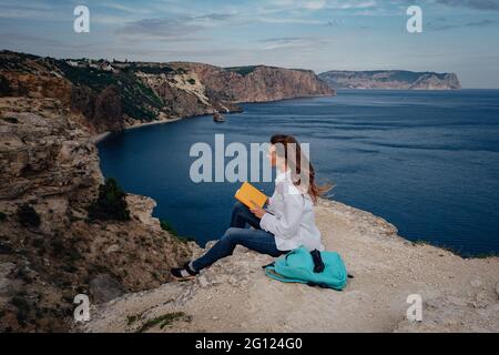 Bella donna è seduta sul bordo della montagna con una vista sul mare, leggendo un libro. Auto-educazione all'aria aperta. Scrittura di note in diario, Foto Stock